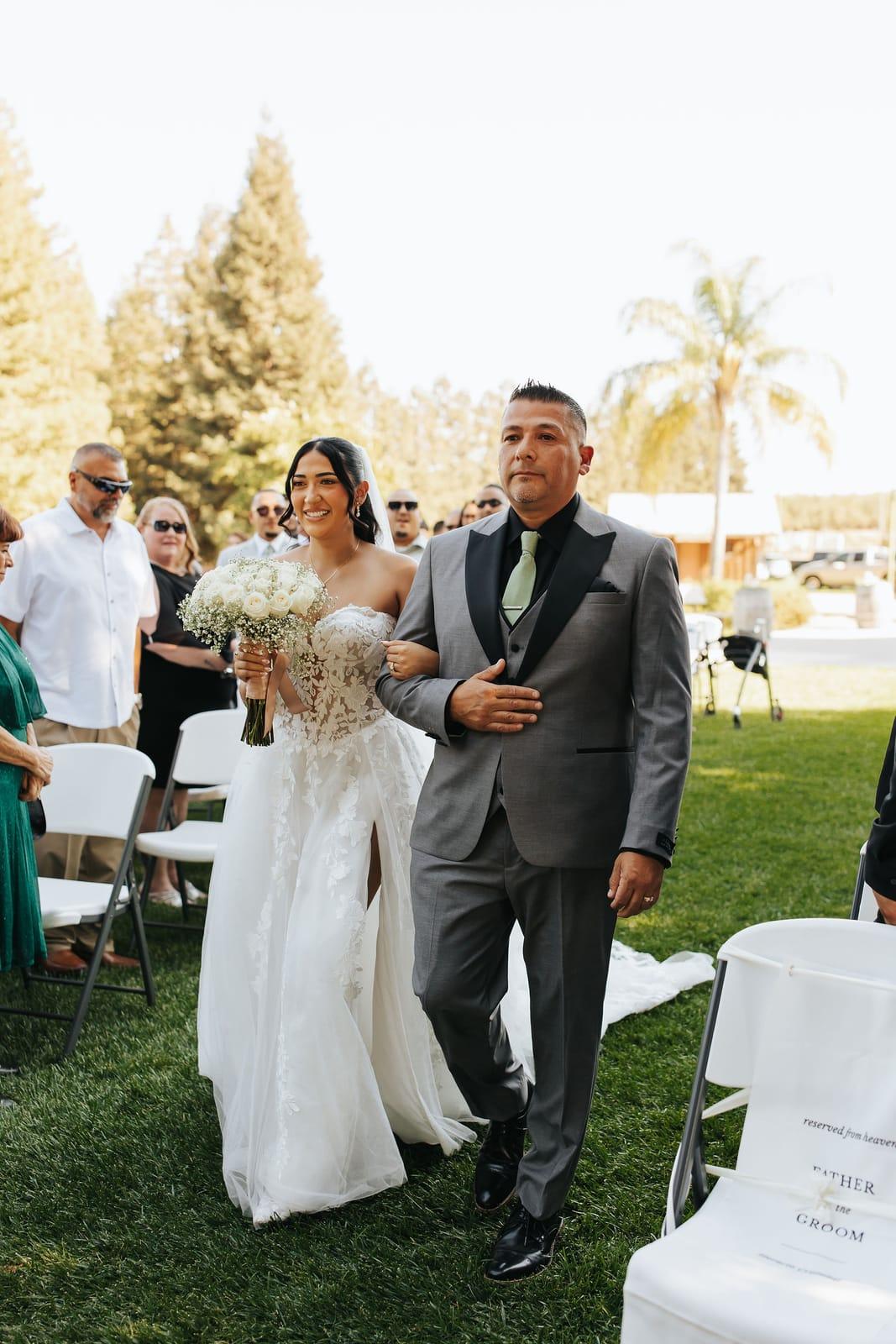 The bride escorted down the grass aisle, bouquet in hand